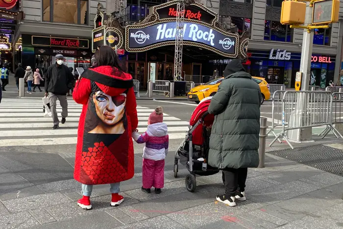 A woman in a bold red coat with a woman's face on the back stand on a Times Square street corner holding a child's hand